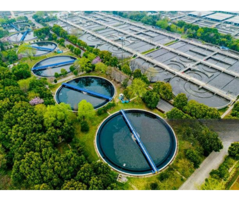 An aerial view of a modern, large-scale sewage treatment plant with multiple circular tanks surrounded by green trees, representing the best STP manufacturers in India.